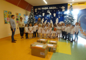 Pani dyrektor Maria Królikowska przekazuje panu Zielińskiemu życzenia świąteczne dla mieszańców hospicjum, Dzieci trzymają kartki świąteczne, na środku paczki z darami.
